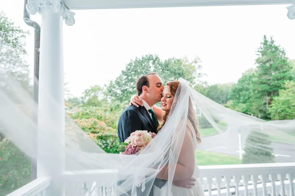 The groom kisses the bride under her veil at the porch railing above the lawn