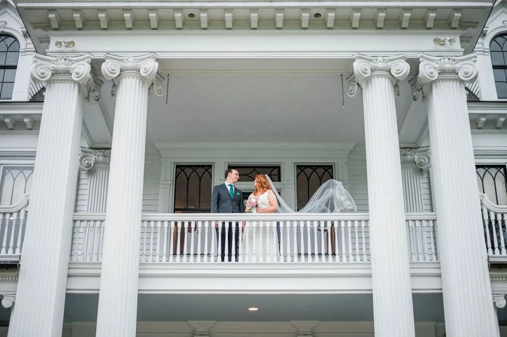 The couple stands on the upper balcony framed between tall white columns