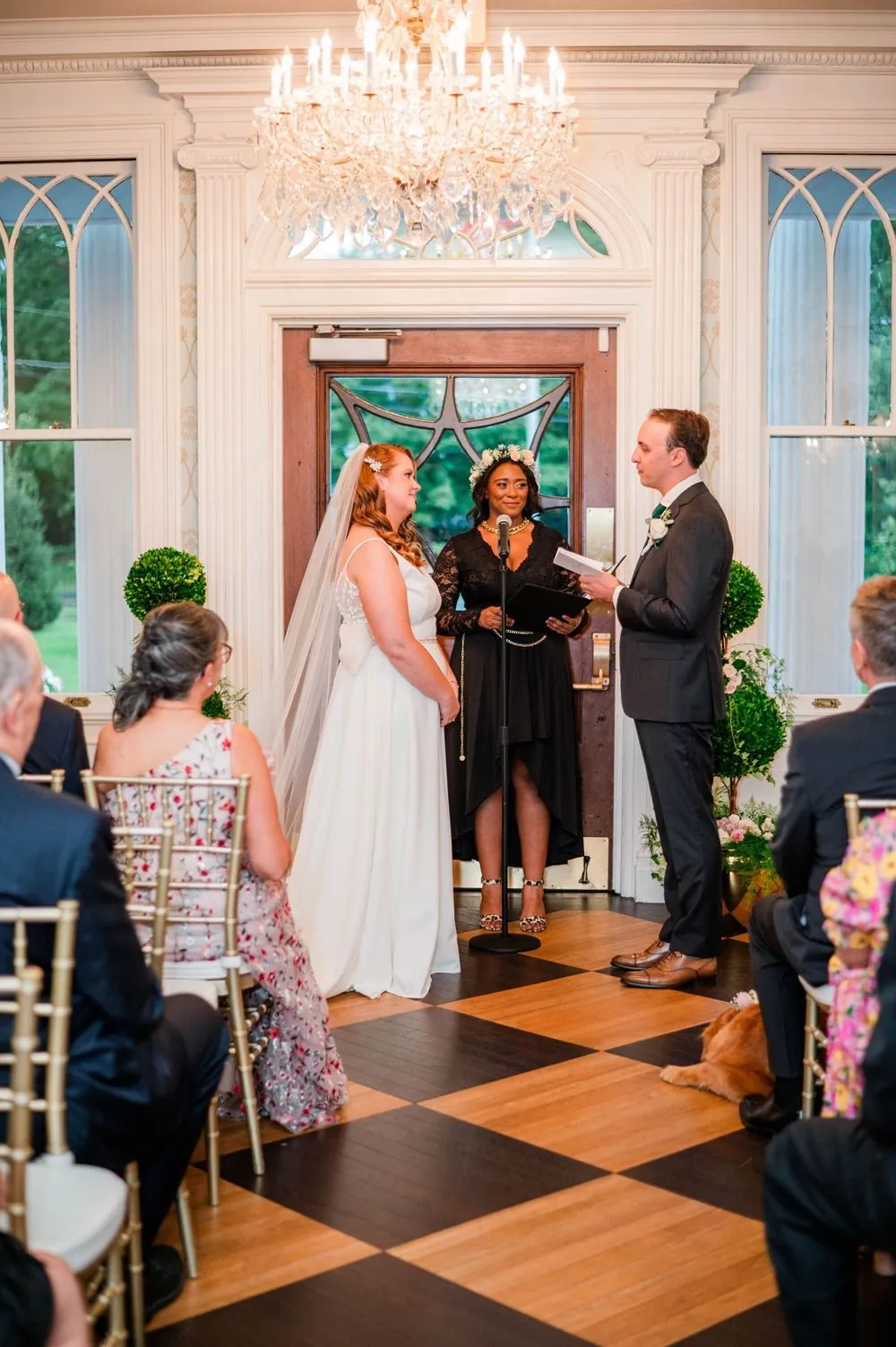 The couple stands before guests in the front parlor under a chandelier