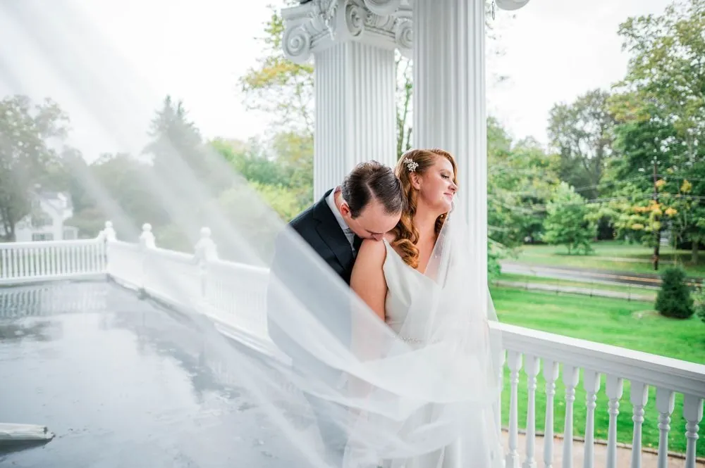 Andrew kisses Maggie's shoulder by a porch column as her veil drifts across the wet floor