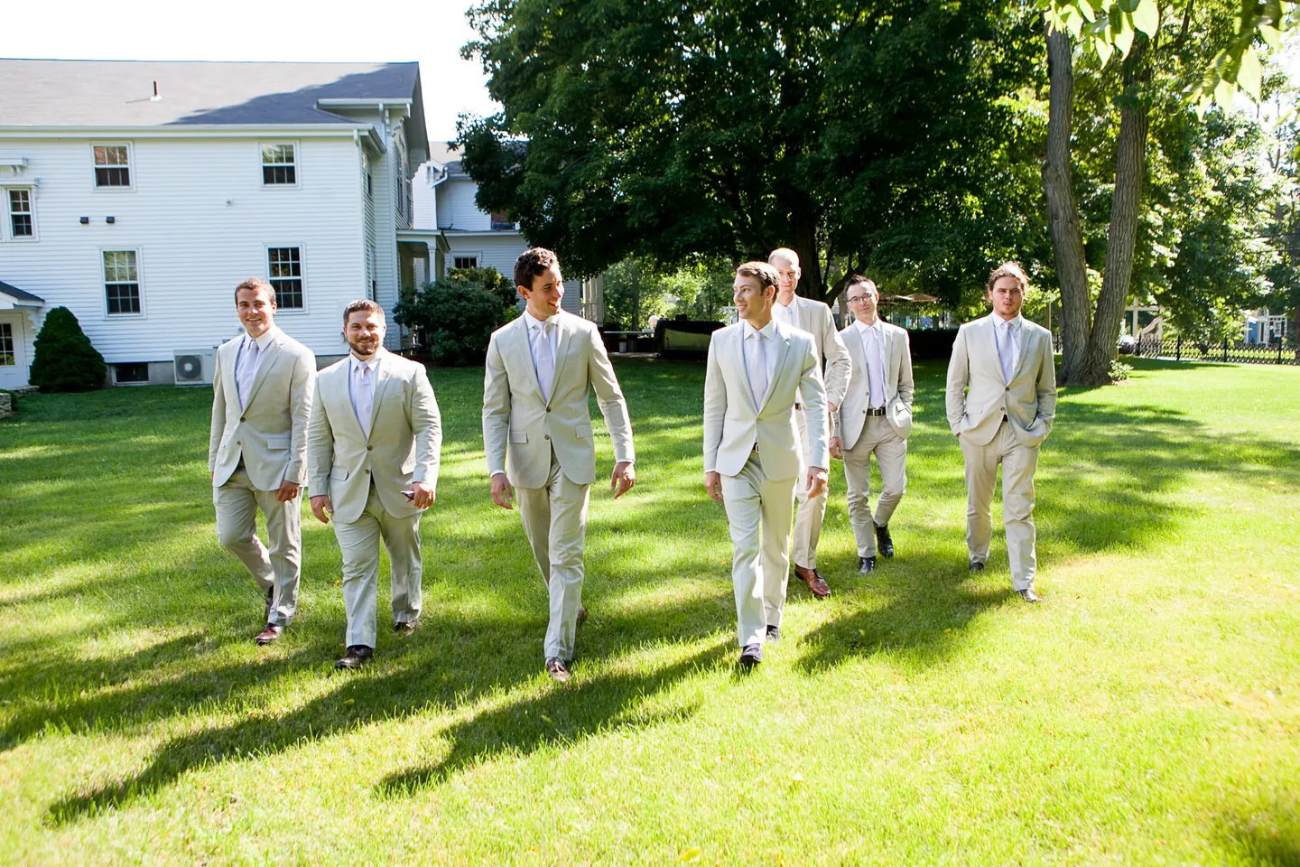 Groomsmen in pale grey suits stride across a wide lawn in front of a white colonial house