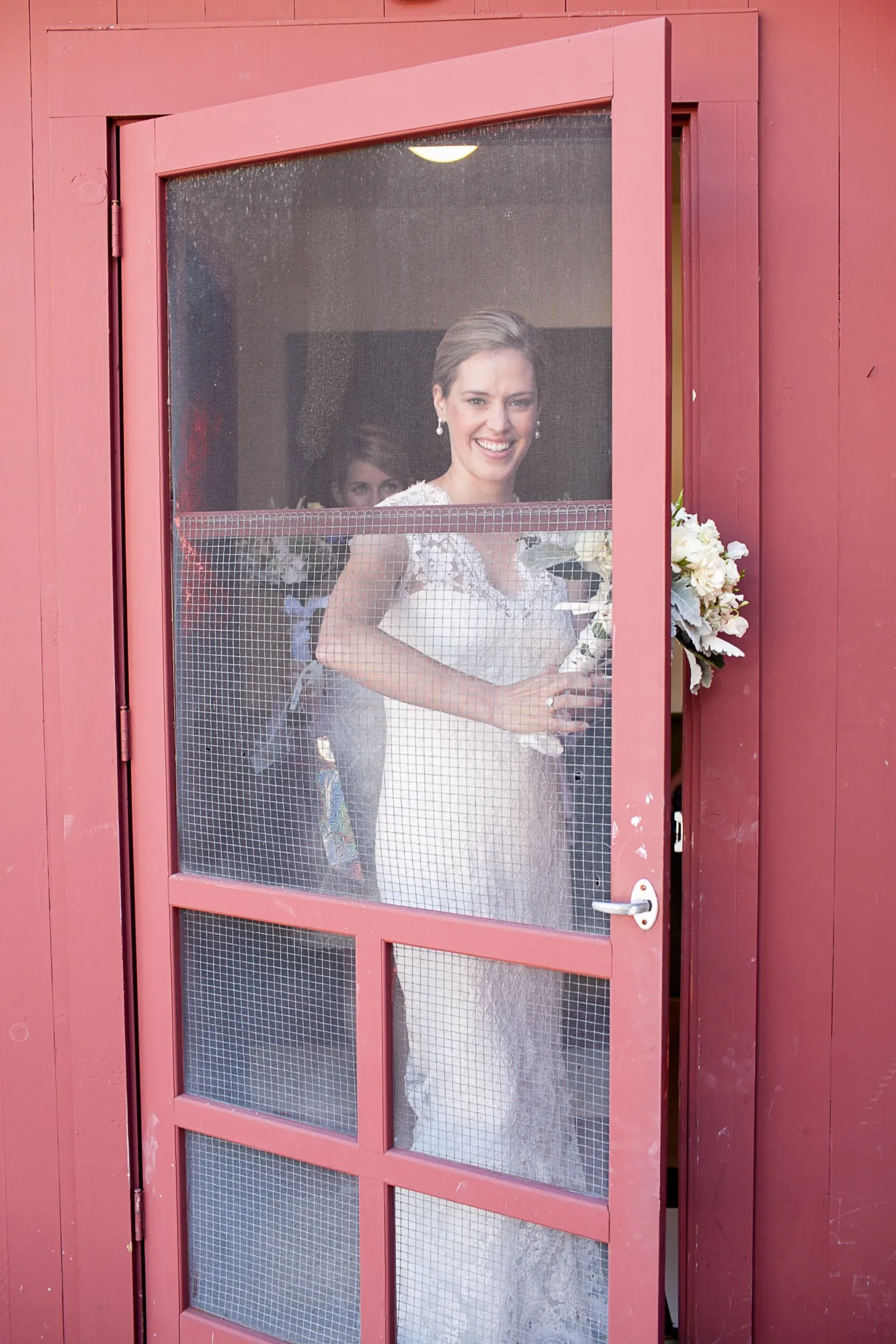 The bride looks out through a red screen door holding her bouquet