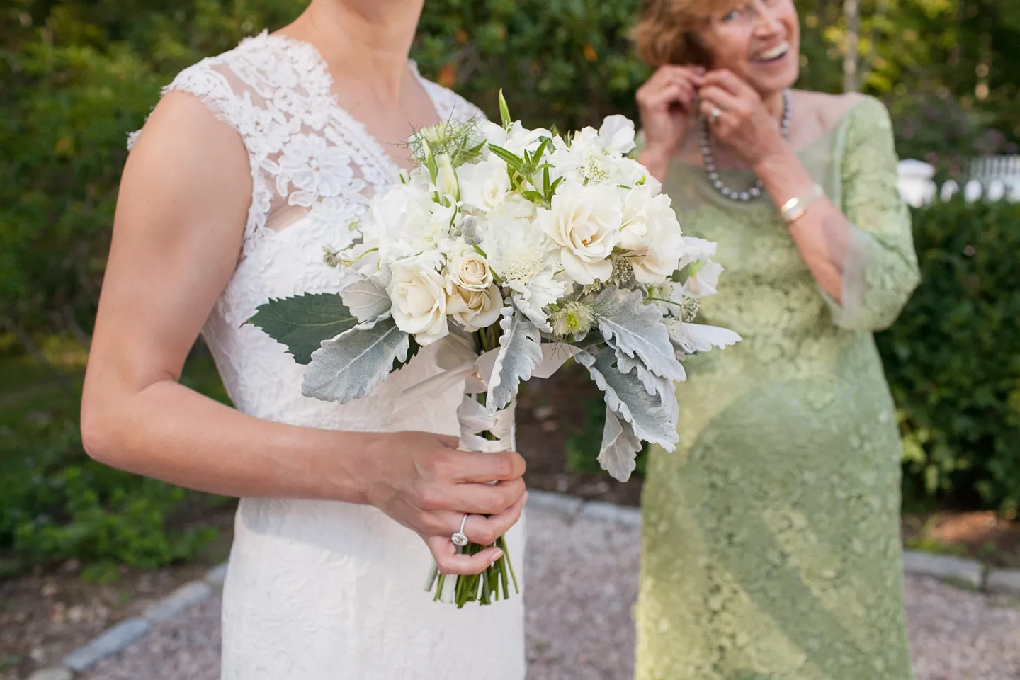 The bride holds a white bouquet with dusty miller beside a woman in a green lace dress
