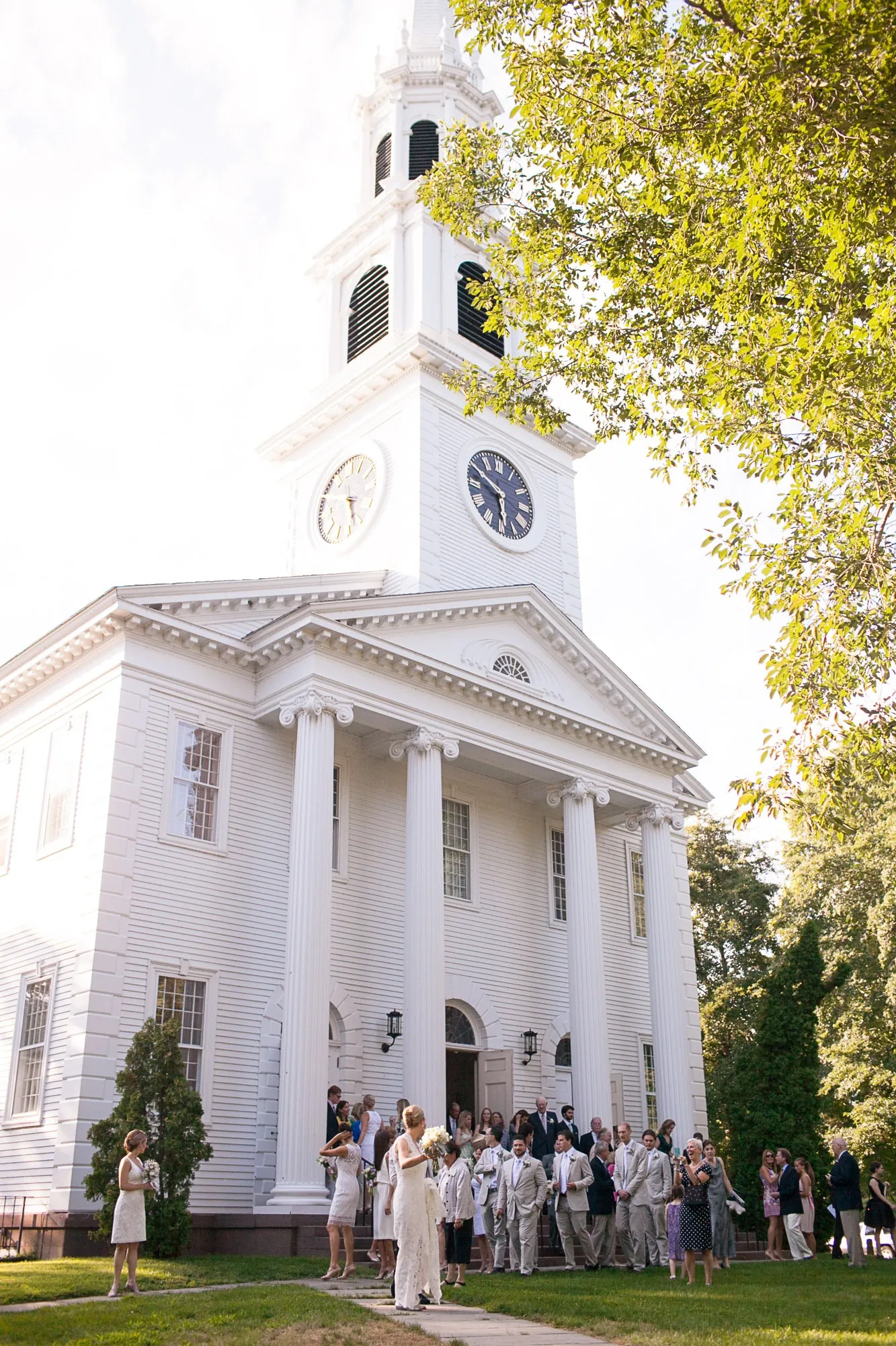 A white church steeple rises above green trees against a pale sky