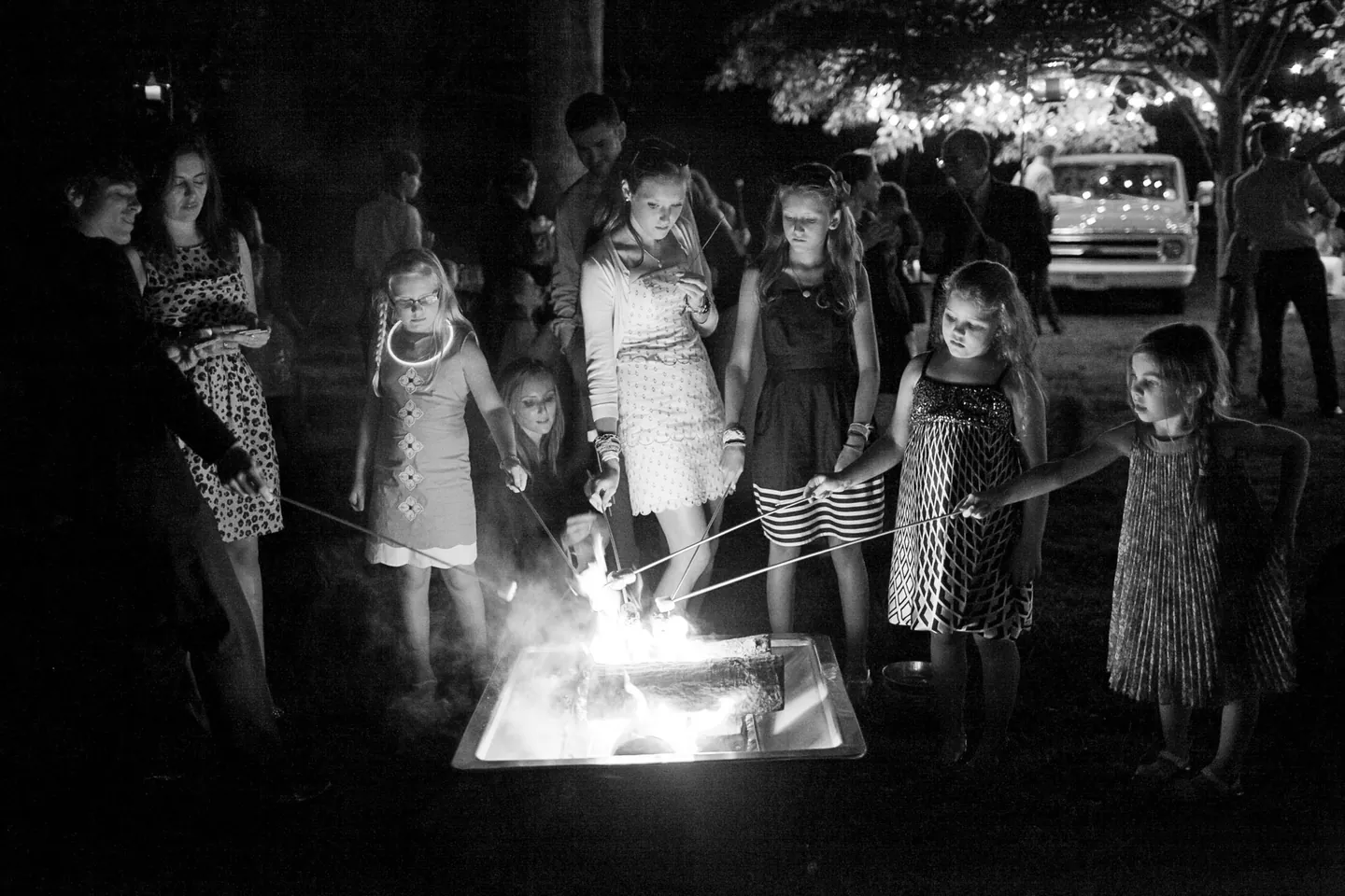 Black and white frame of kids roasting marshmallows over a fire pit at night