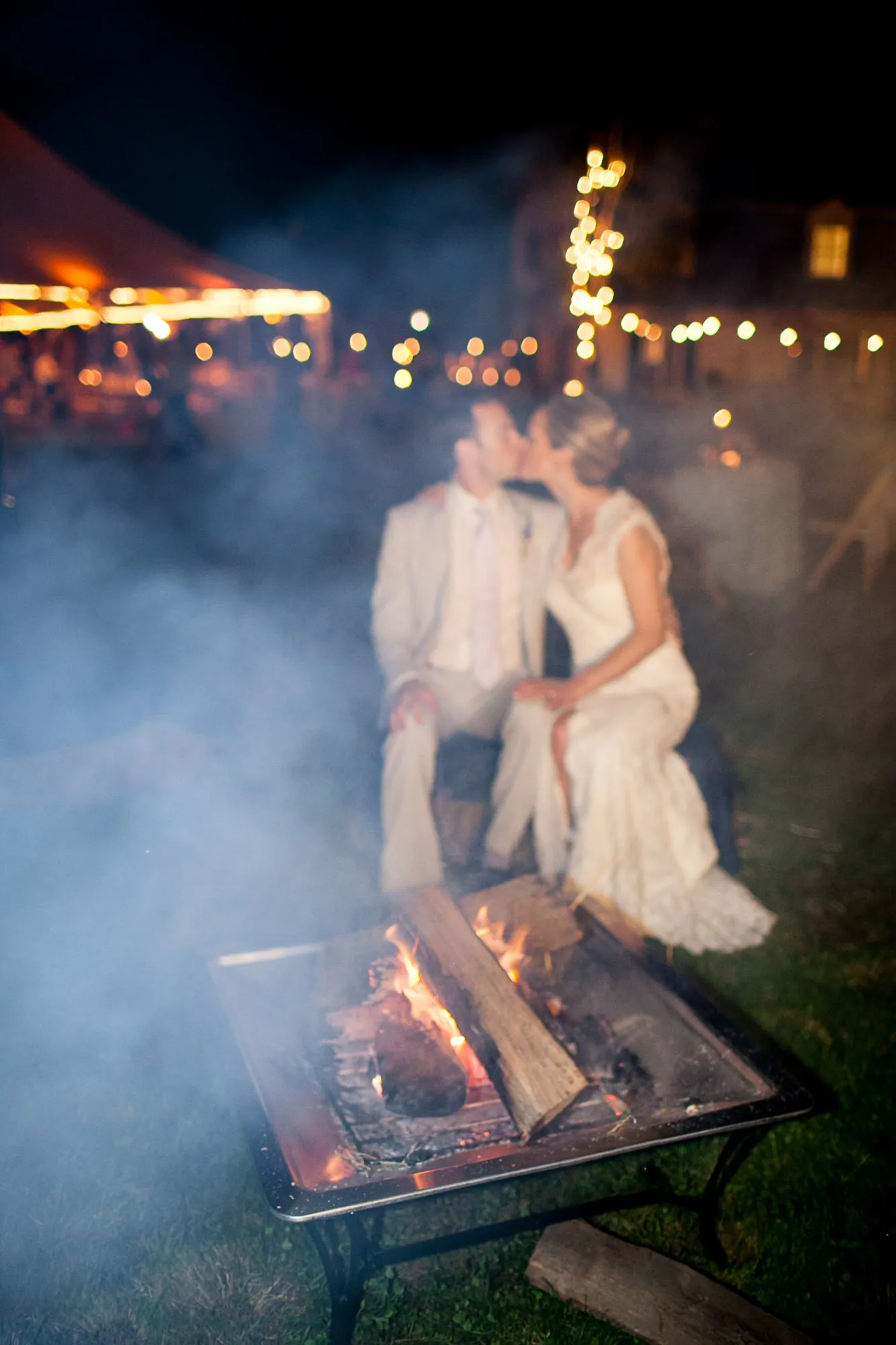 Marina and James kiss beside a fire pit as smoke drifts toward the string lights