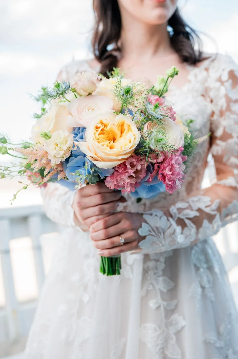 Bride holds a loose bouquet of peach roses and pale blue flowers against her long lace sleeves
