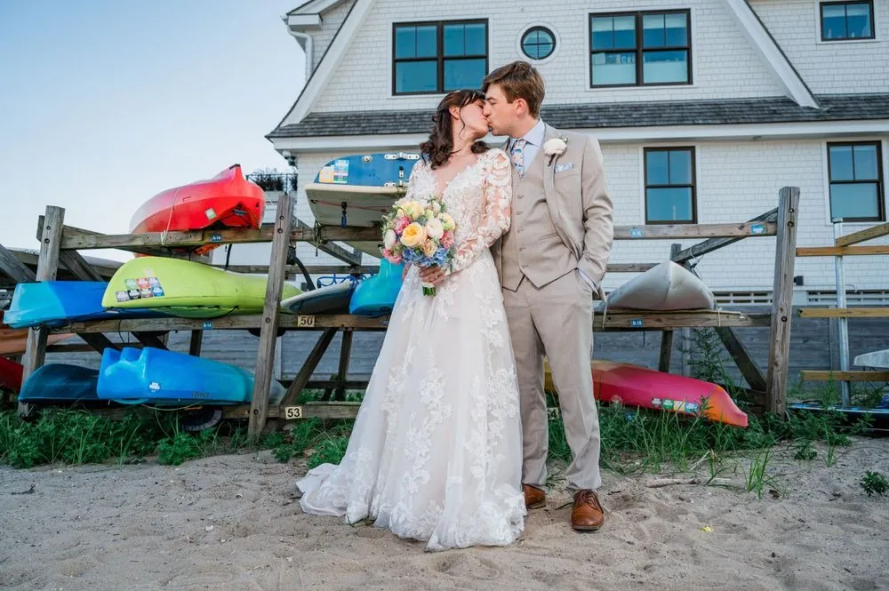 Mary and Dan kiss in front of wooden racks stacked with red, yellow, and teal kayaks