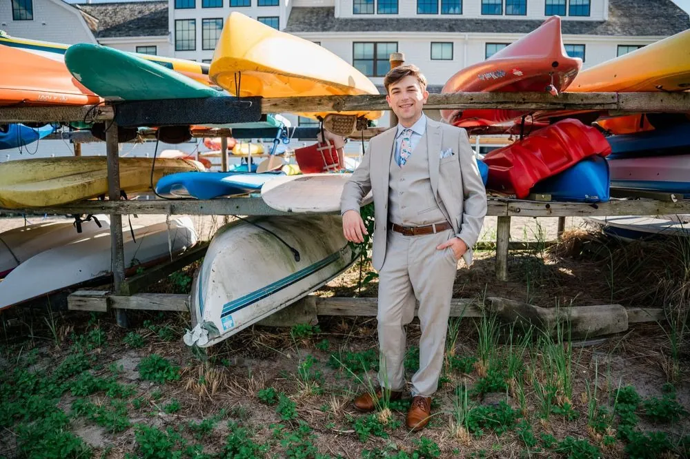 Groom stands with hands in pockets in front of a rack of colorful kayaks and beach grass