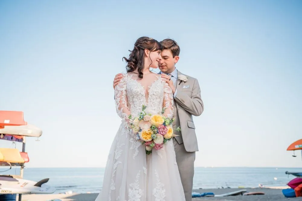 Dan kisses Mary's forehead as she smiles holding her pastel bouquet on the beach