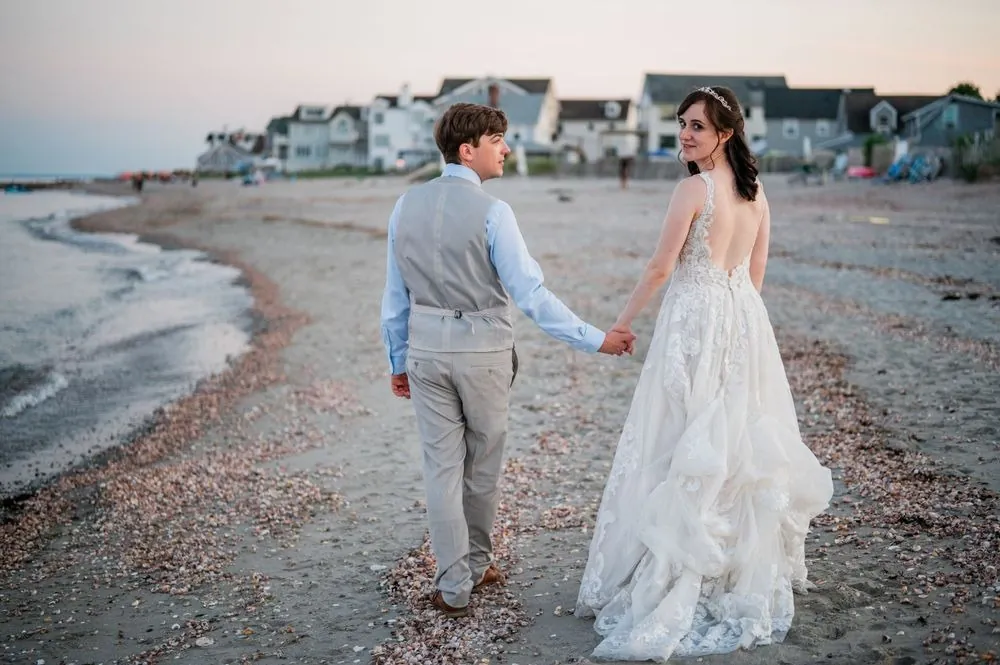 Mary and Dan walk hand in hand along a curving shell beach with houses behind