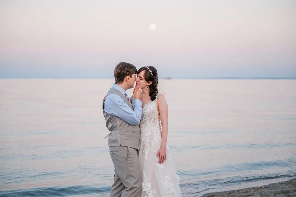 Mary and Dan touch foreheads beside flat pastel water with a small moon above the horizon