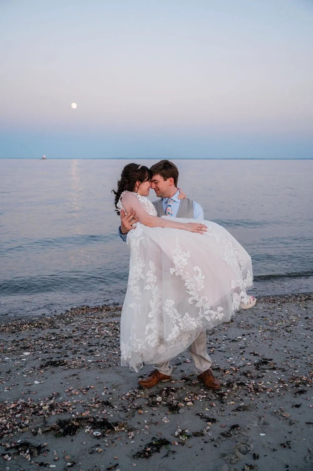 Dan lifts Mary off the sand at the tide line under a pale evening moon