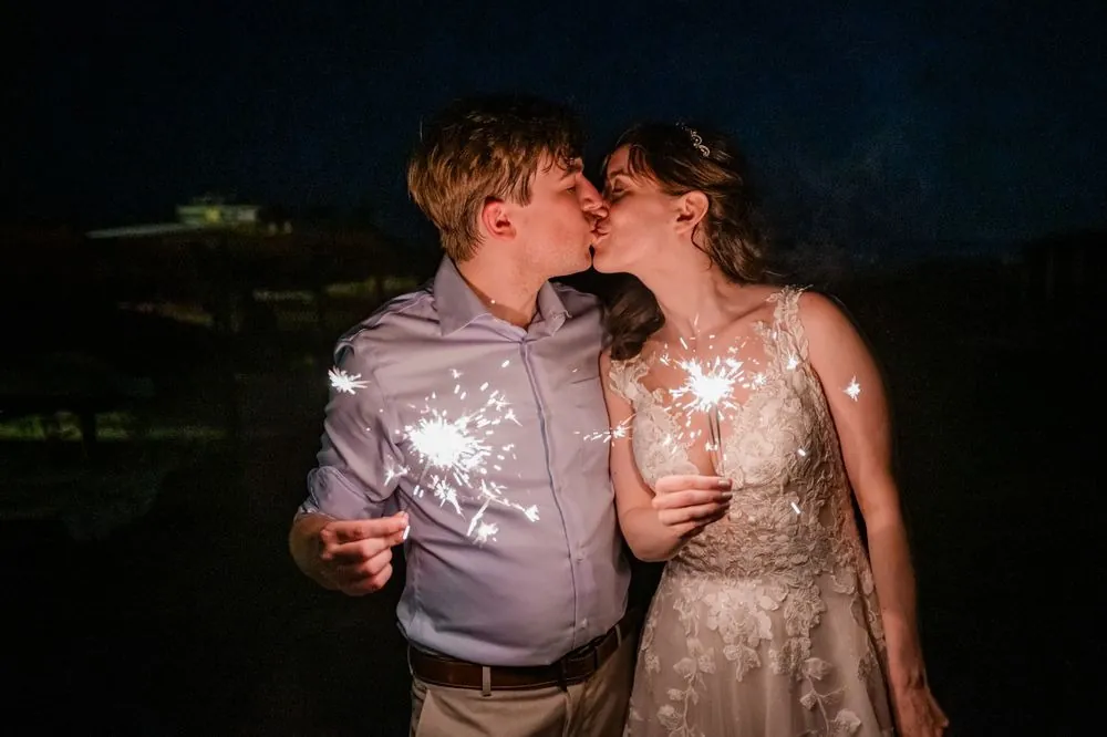Mary and Dan kiss holding lit sparklers against the dark night sky