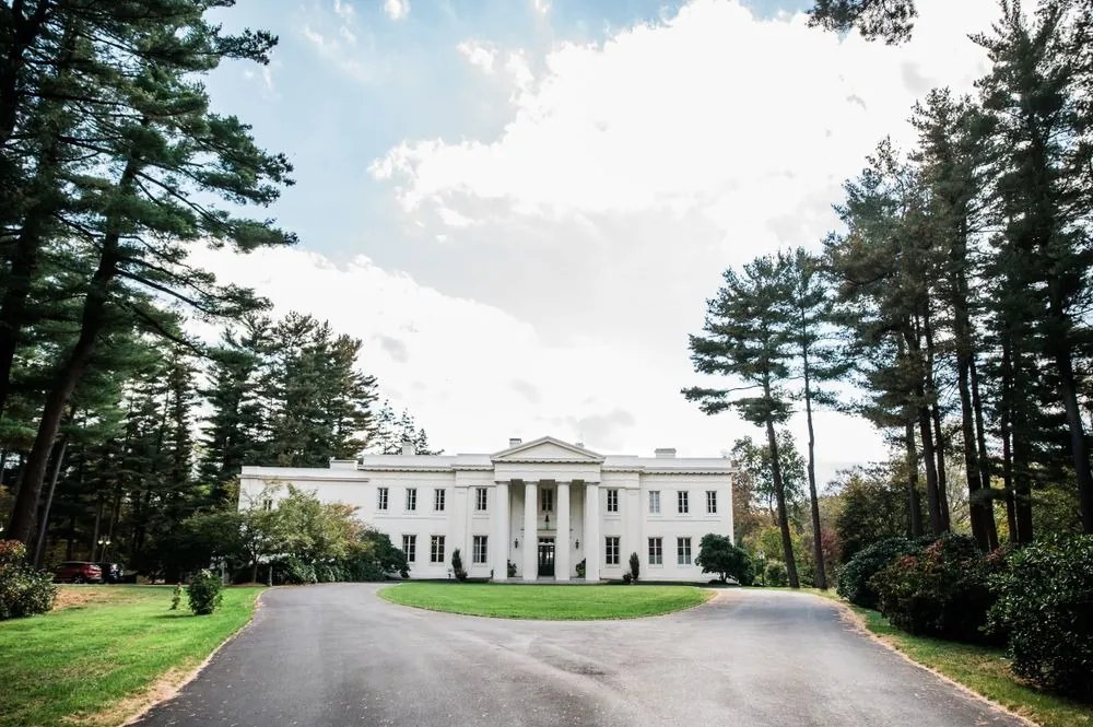 The white mansion at the end of a long drive lined with tall pines under a blue sky