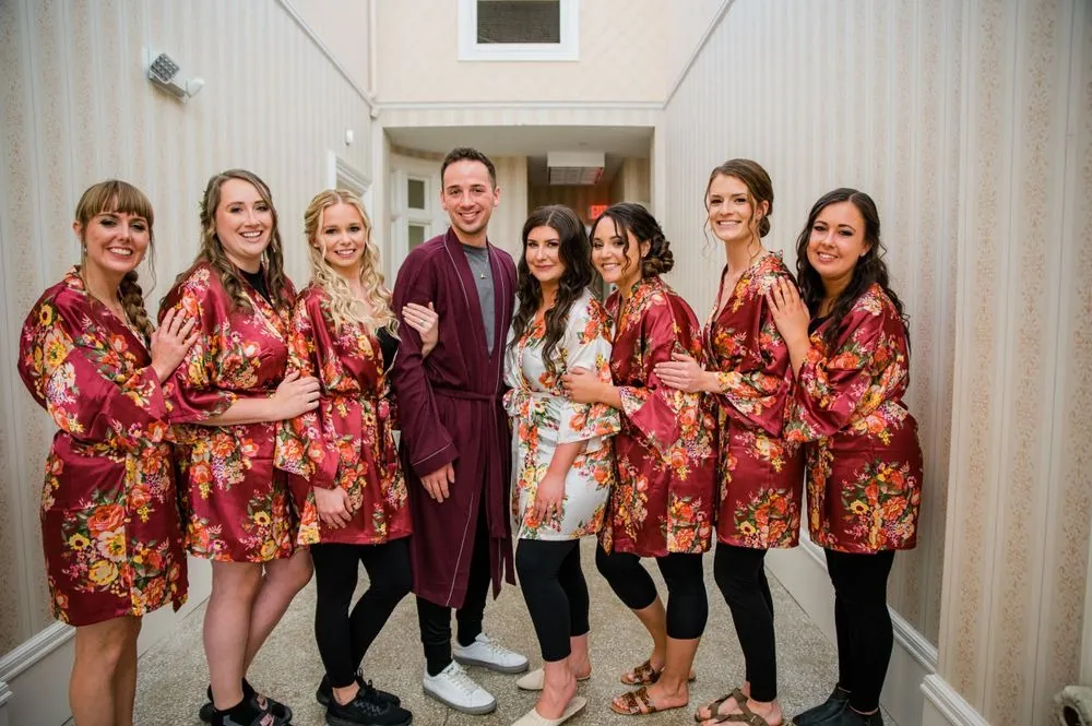 Bridesmaids in matching red floral robes laugh together in a white hallway