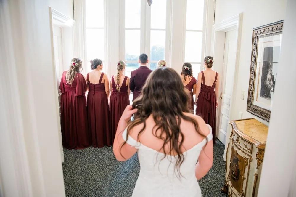 The bride steps toward her bridesmaids in burgundy gowns as they turn to see her