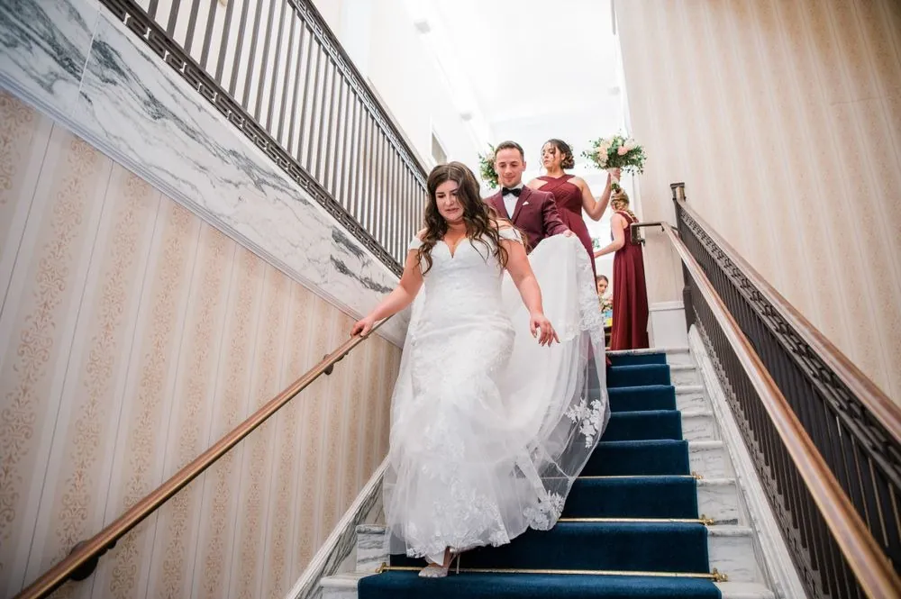 The bride walks down a carpeted staircase holding her skirt, bridesmaids behind her