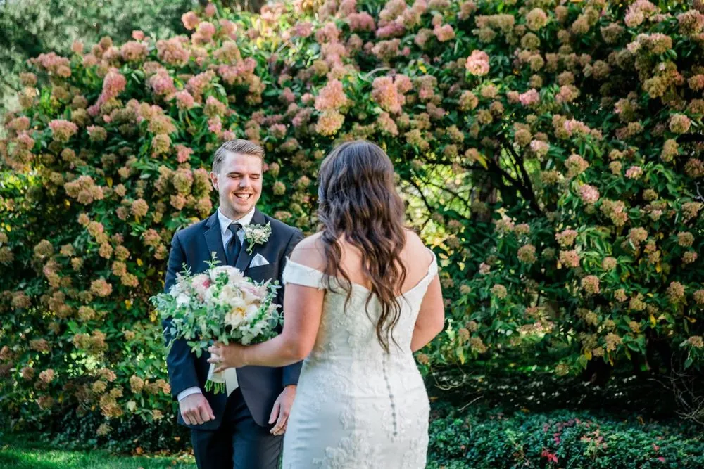 The groom turns to see the bride in front of a hedge of fall hydrangeas