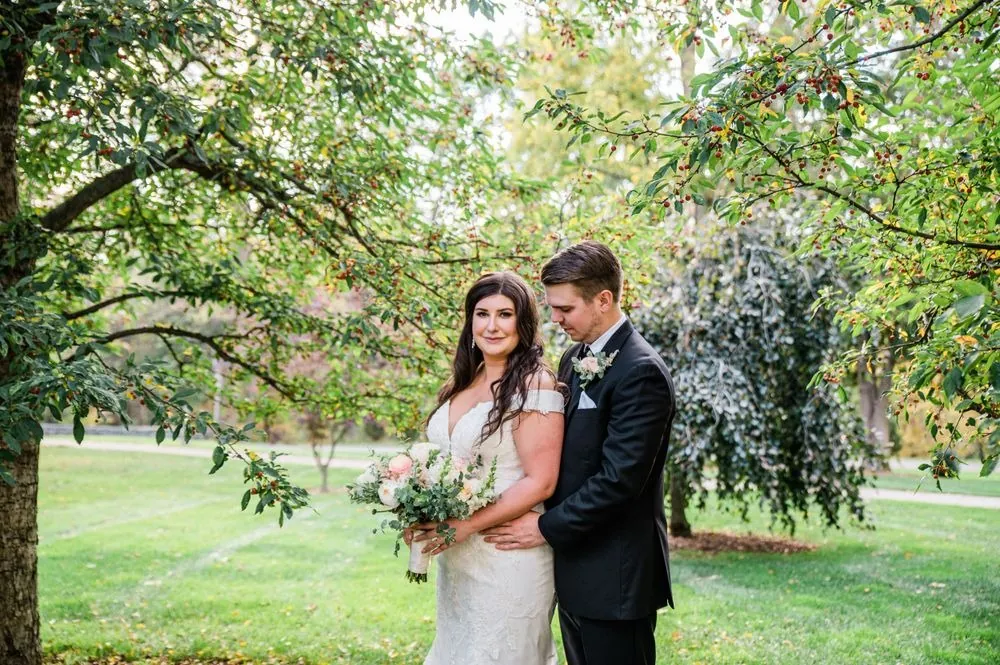 The bride and groom stand under a spreading tree on the green lawn