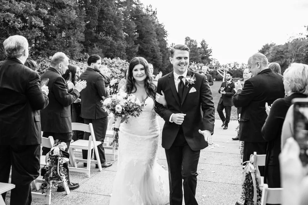 Black and white frame of the newlyweds walking back up the aisle among guests