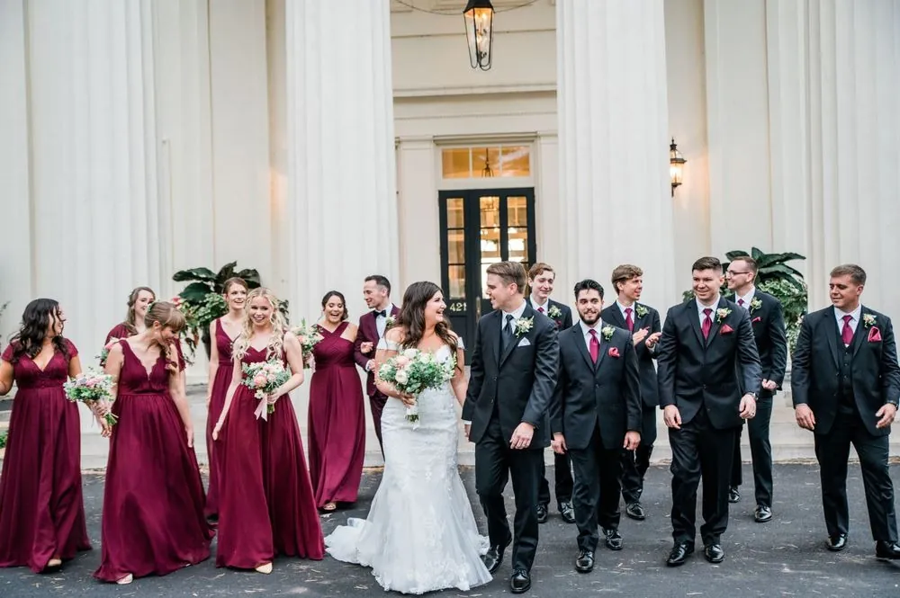 The wedding party walks in pairs toward the camera beside the white columns
