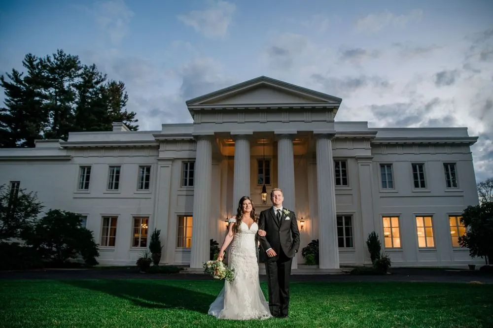 Megan and Jacob stand on the lawn before the lit mansion portico at dusk