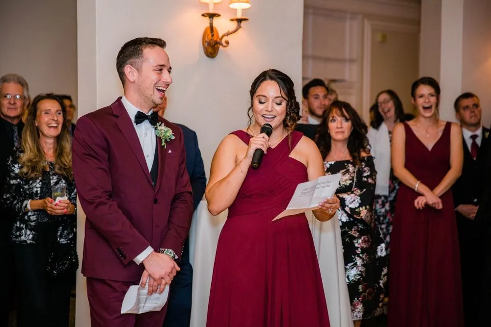 A bridesmaid in burgundy gives a toast while a groomsman in a burgundy jacket laughs