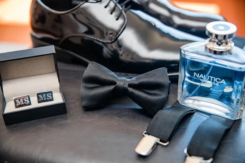 Groom's black shoes, bow tie, initialed cufflinks, and cologne arranged on dark leather