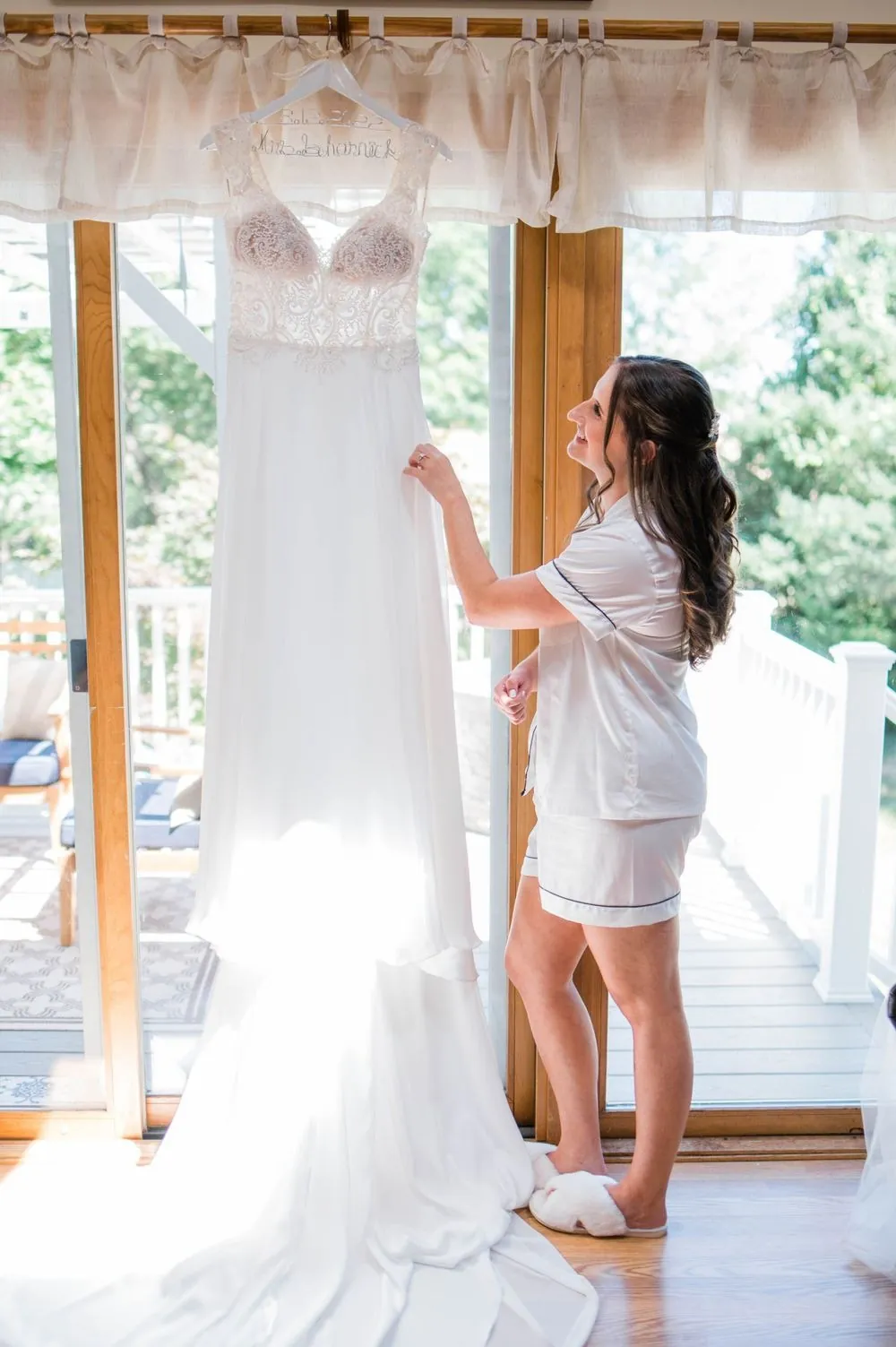 Bride in a robe touches her gown hanging in a bright doorway