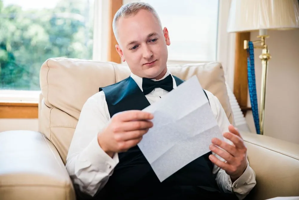 Groom in a black vest and bow tie reads a letter in a cream armchair