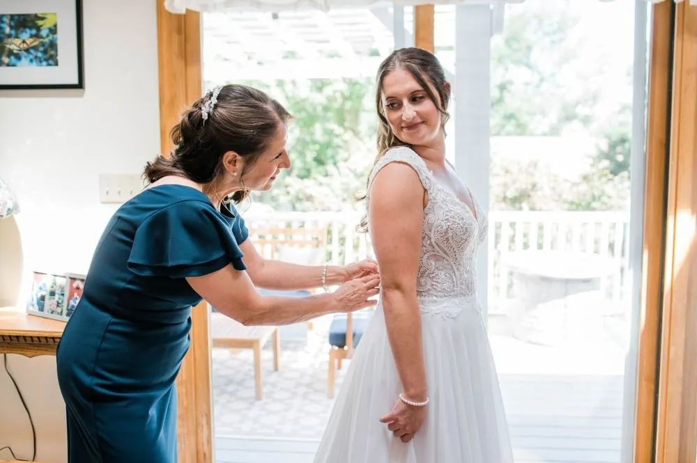 Woman in a teal dress fastens the back of the bride's gown in a sunny doorway