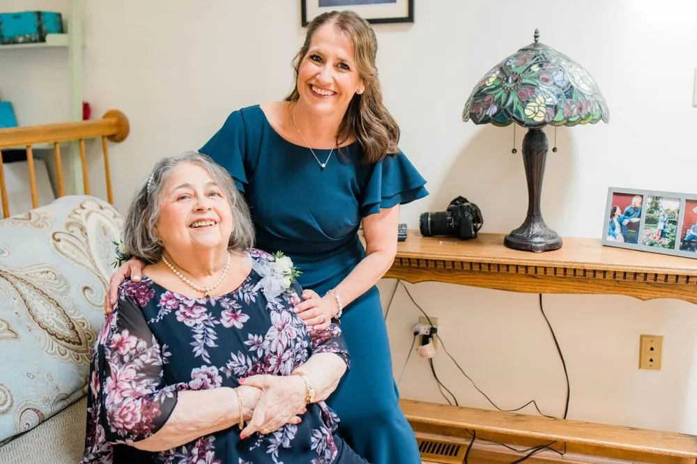 Grandmother in a navy floral dress laughs as a woman in teal hugs her shoulders