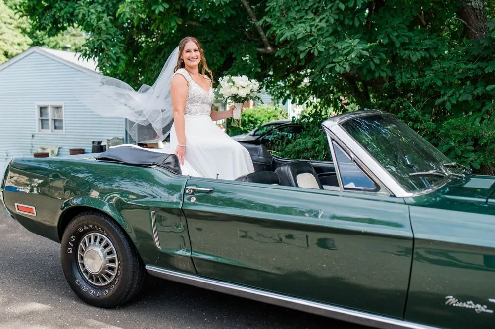 Bride sits on the back of a dark green classic convertible as her veil lifts in the breeze