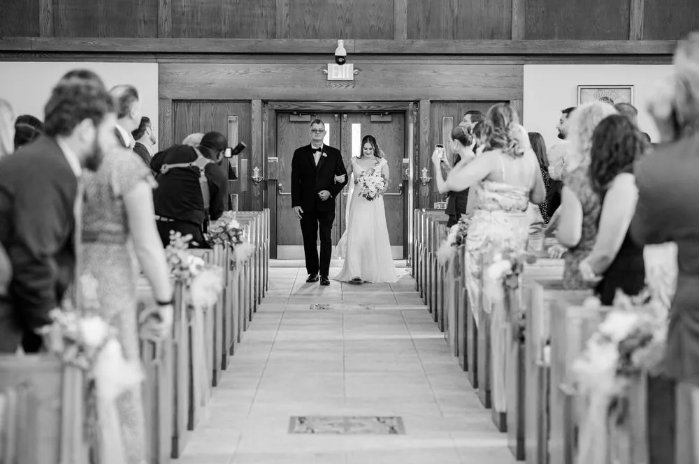 Black and white frame of the bride walking the long church aisle between packed pews