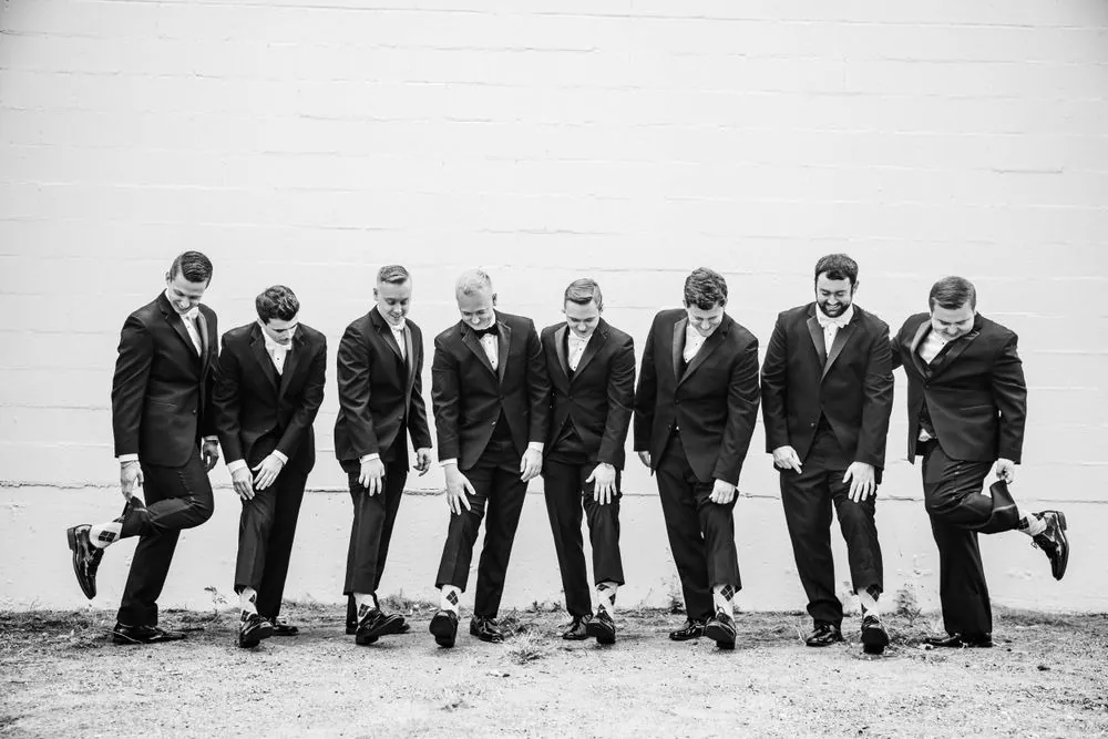 Black and white frame of groomsmen kicking up their heels against a white brick wall
