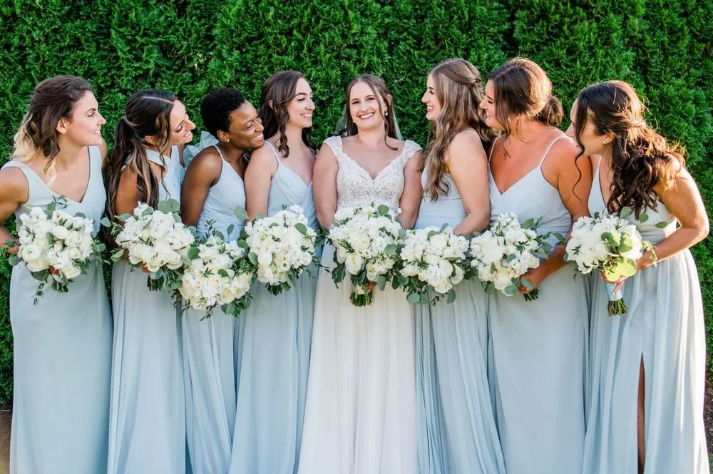 Bridesmaids in dusty blue gowns laugh with the bride against a green hedge