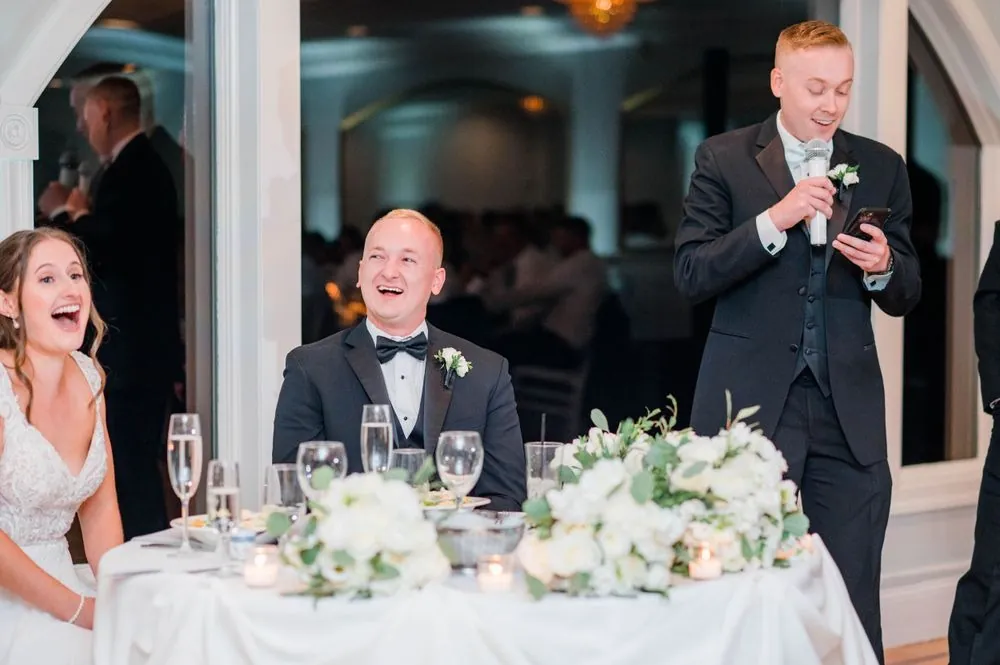 Groom laughs at the head table as a guest in a tuxedo gives a toast into a microphone