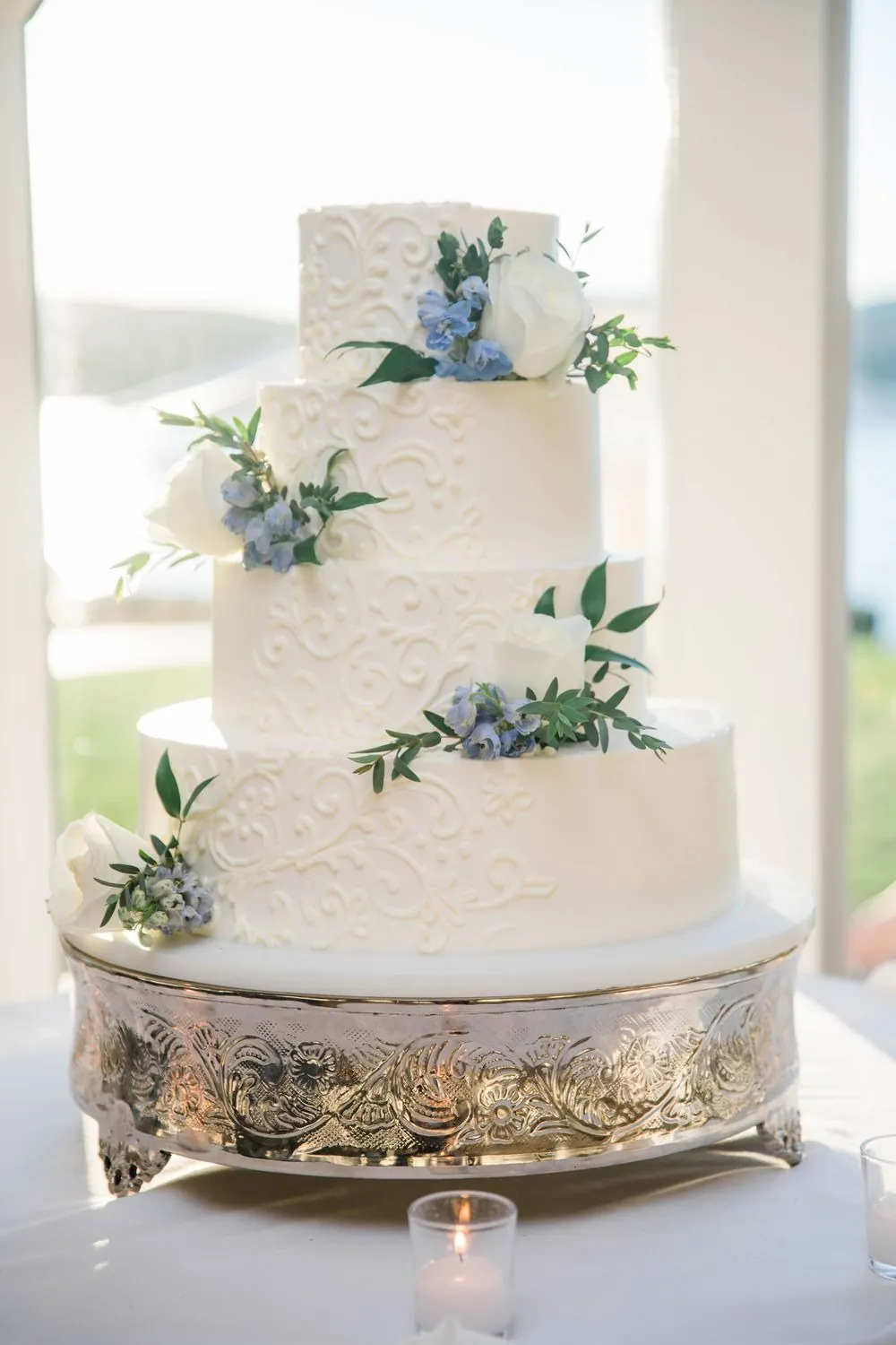 Tiered white cake trimmed with white flowers and blue accents on a silver stand