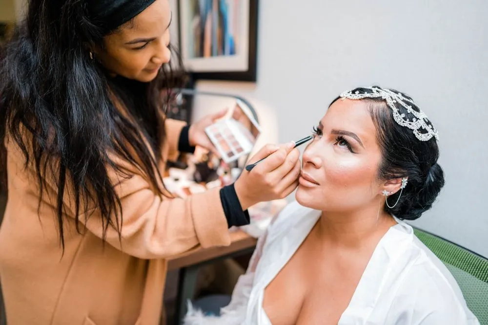 A makeup artist works on the bride, who wears a jeweled headpiece and white robe