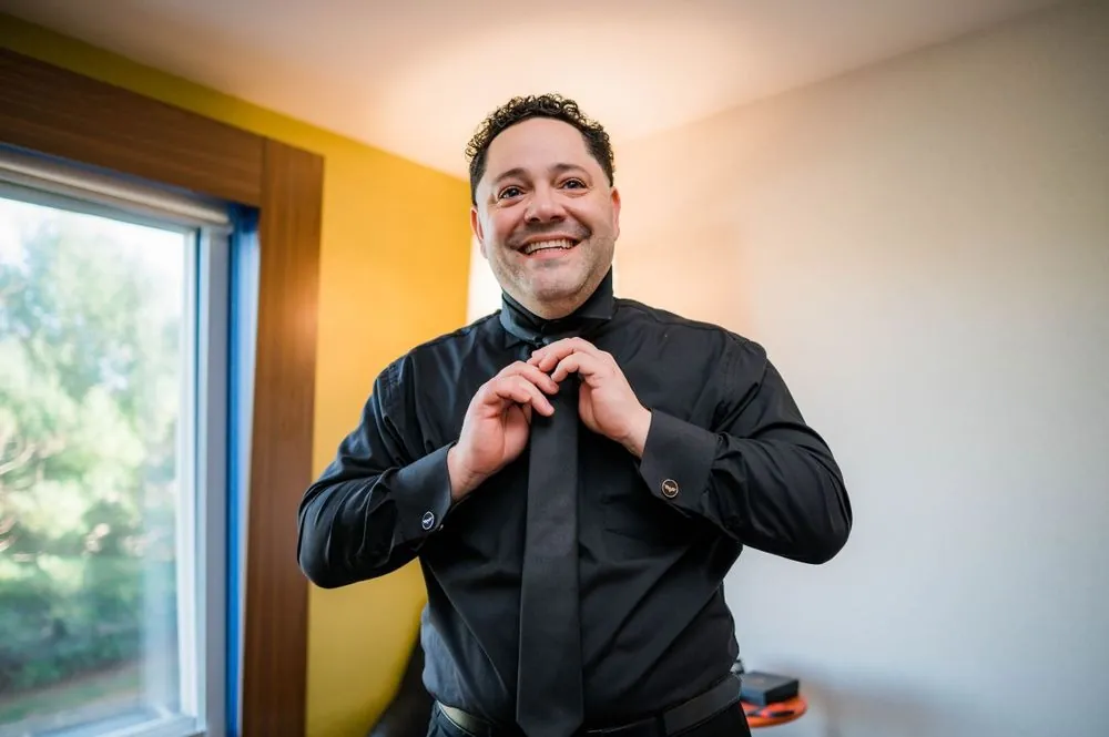 The groom grins as he knots a black tie over a black shirt