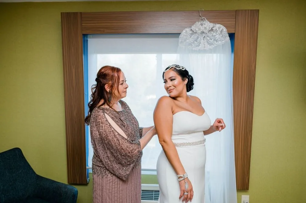 A family member helps the bride into her white gown by the window