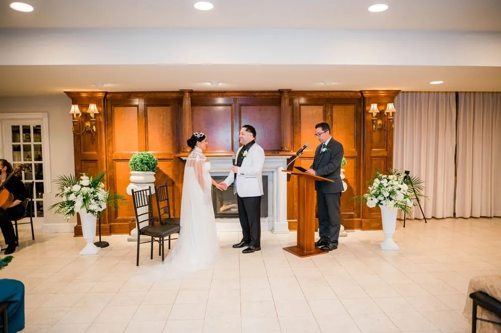 The couple stands before the officiant at a wood-paneled fireplace with white flowers