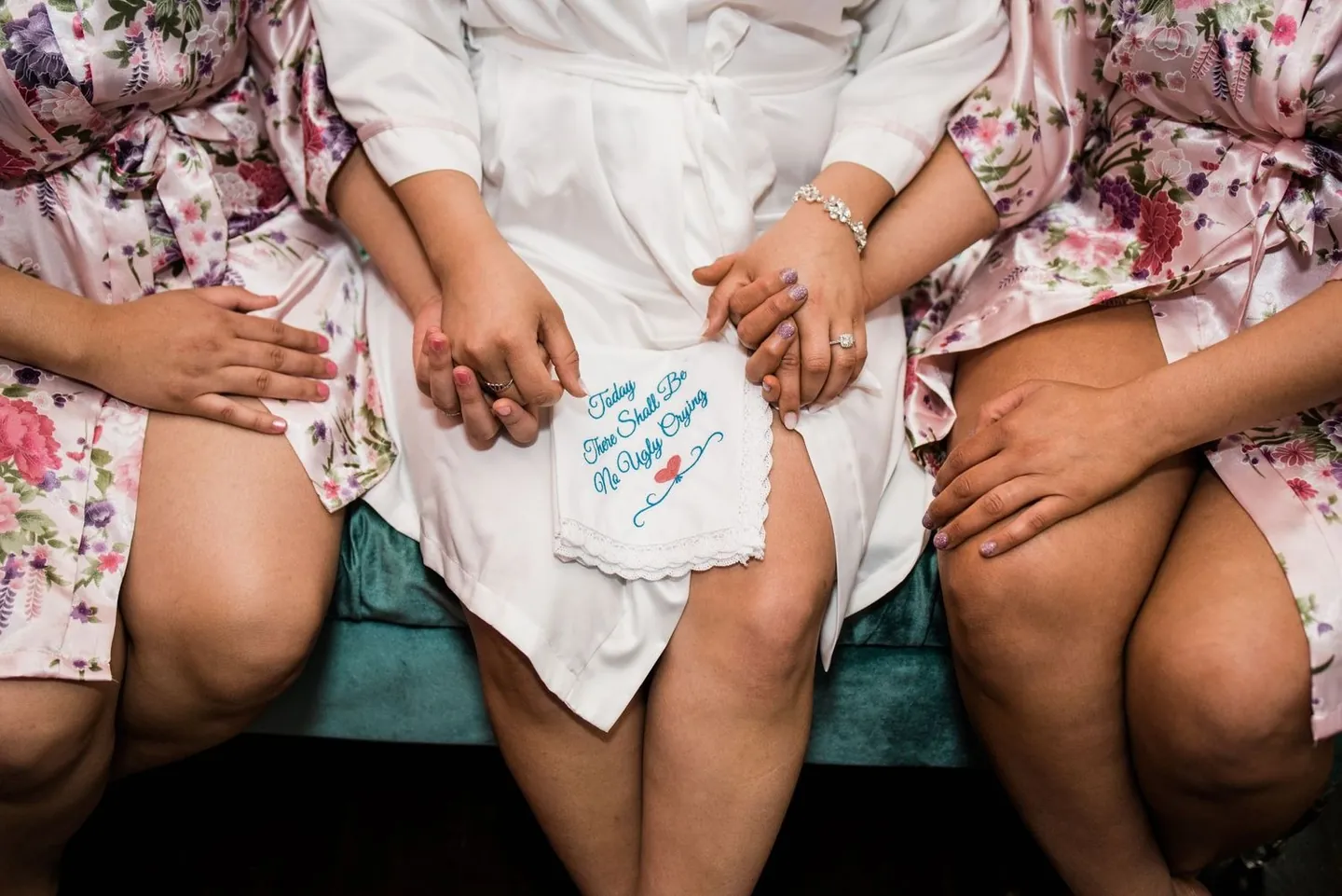 Bridesmaids in floral robes hold the bride's hands over an embroidered handkerchief in her lap