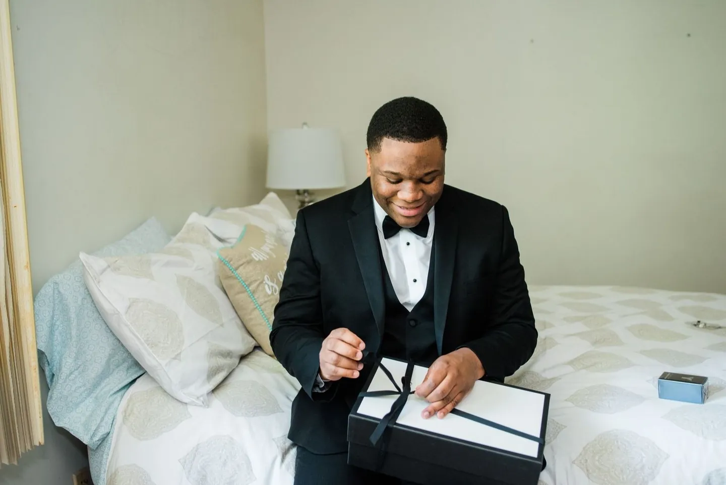 Groom in a black tuxedo opens a gift box while sitting on the edge of a bed