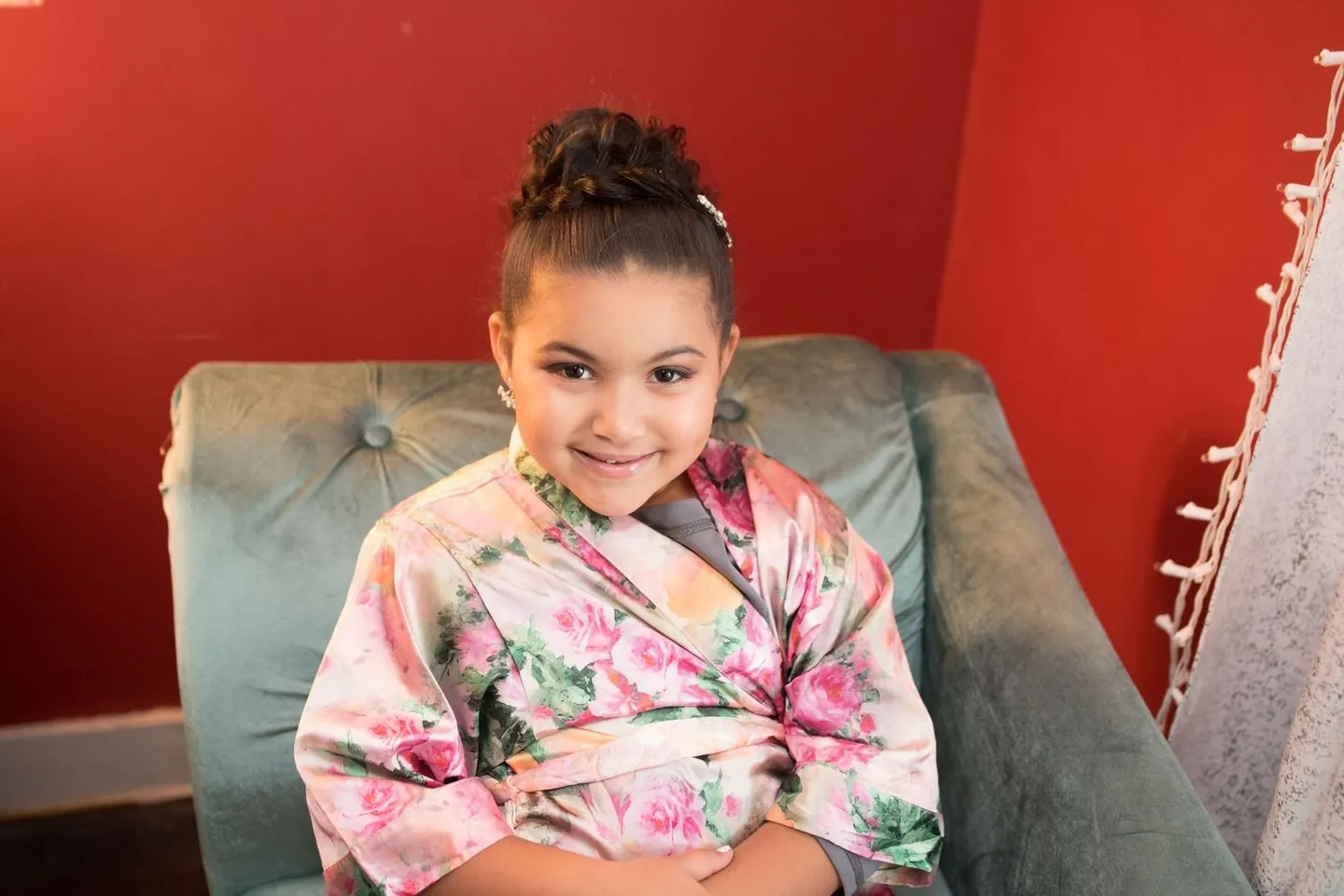 Flower girl in a pink floral robe grins from a teal chair against a red wall