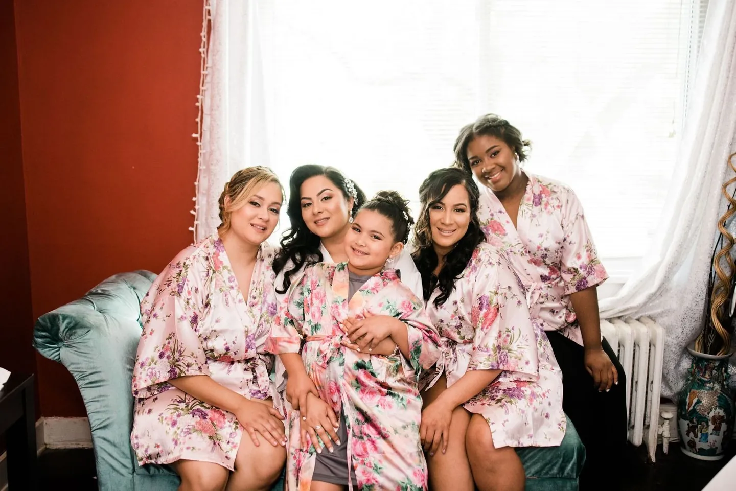Bride in a white robe laughs with bridesmaids in pink floral robes on a sofa