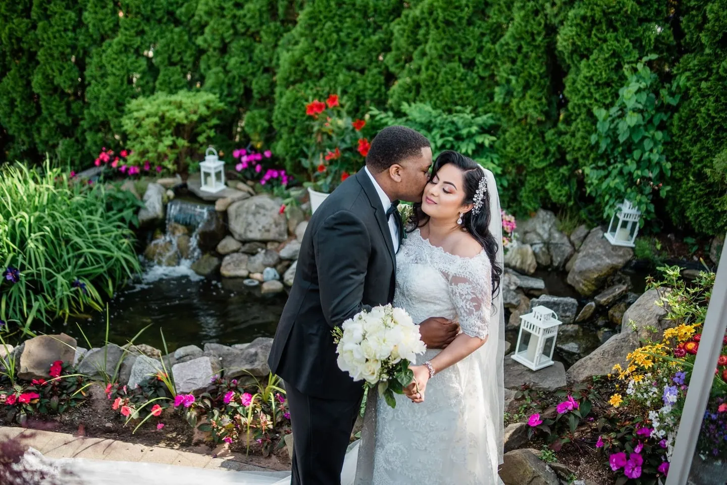 Jordan kisses Natasha's cheek beside a rock waterfall and bright garden flowers