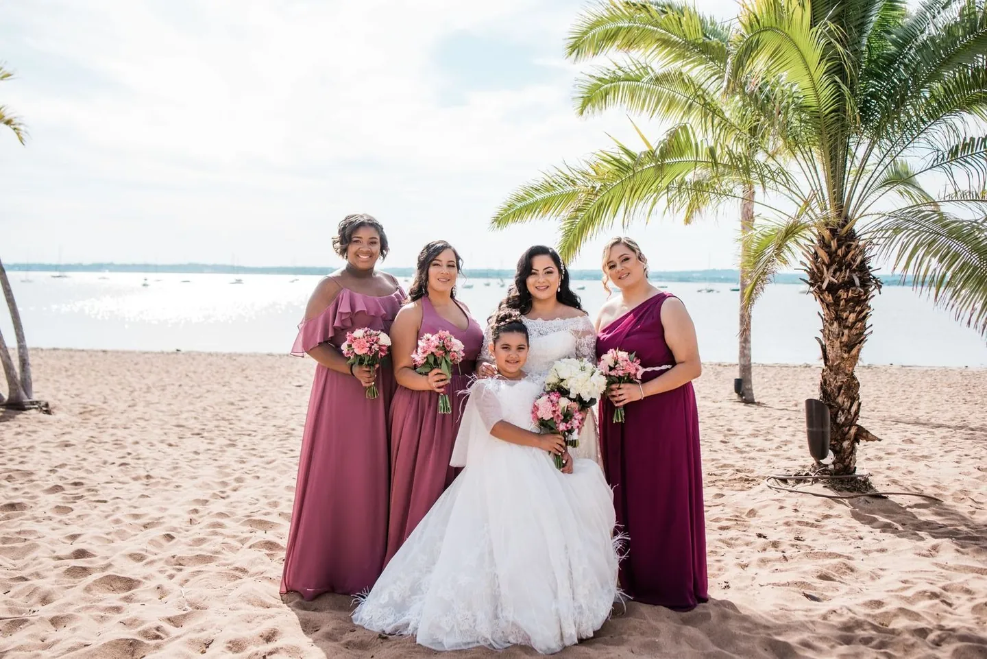 Bride stands with bridesmaids in plum gowns under palm trees on the beach