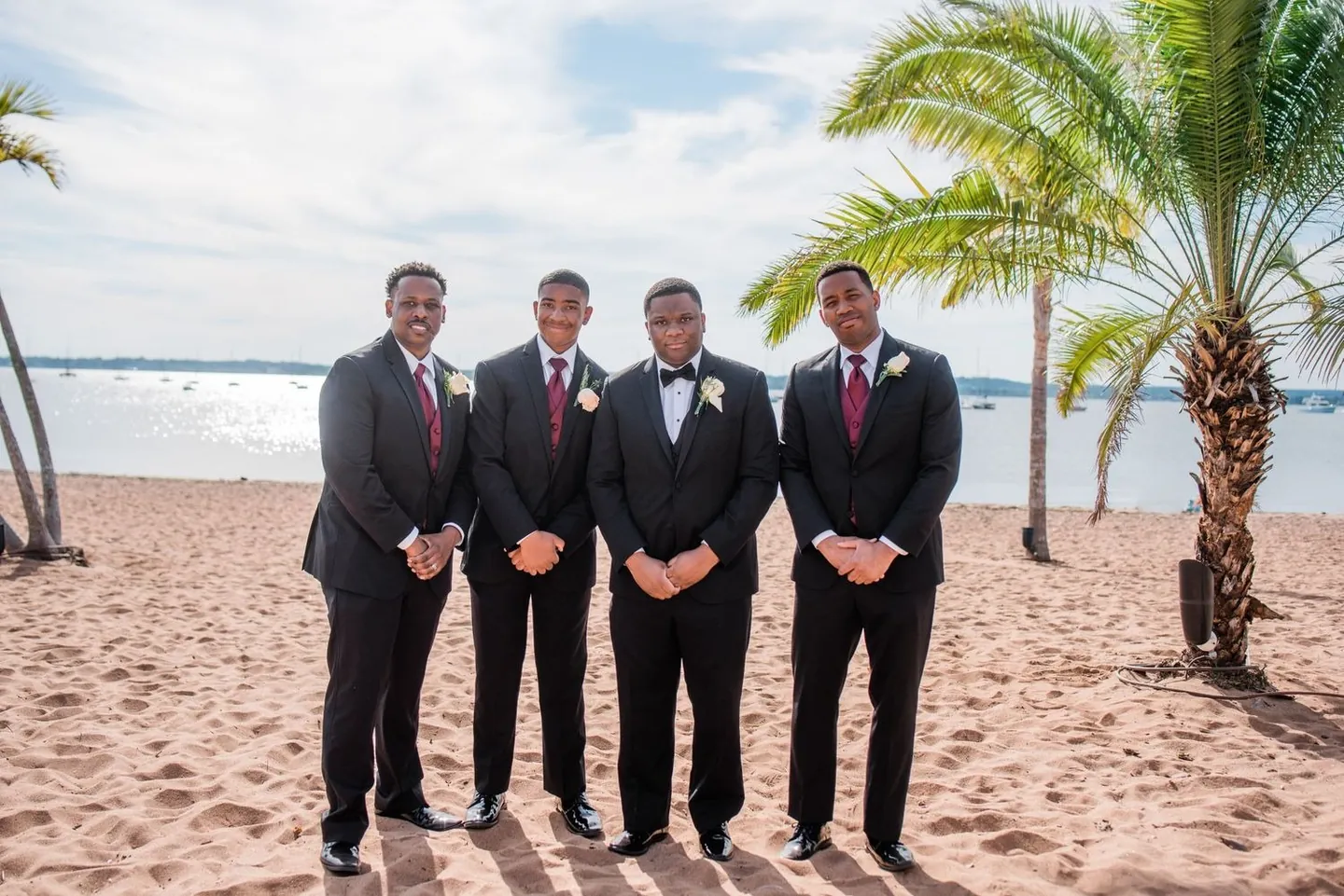 Groom and groomsmen in black tuxedos and burgundy ties stand on the sand by palm trees
